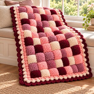 May include: A crochet blanket featuring a textured bubble stitch pattern in a checkerboard arrangement of cream, light pink, mauve, and deep burgundy squares. The blanket is finished with a scalloped border of cream and burgundy yarn.