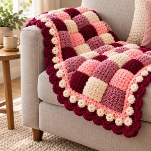 May include: A handmade crochet blanket featuring a checkered pattern of cream, light pink, mauve, and deep burgundy squares. The edges are finished with a scalloped border accented by small, cream-colored crocheted flower motifs.