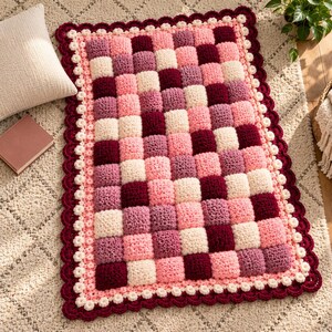 May include: A rectangular crochet rug featuring a checkered pattern of raised, puffy squares in shades of cream, light pink, mauve, and deep burgundy. The rug is finished with a decorative scalloped border in cream and dark burgundy.