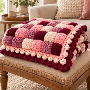 May include: A crochet blanket featuring a checkered pattern of square blocks in shades of cream, light pink, mauve, and deep burgundy. The blanket is finished with a decorative scalloped border composed of small cream-colored flower motifs.