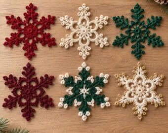 Puede incluir: Seis adornos de copo de nieve tejidos a ganchillo dispuestos en dos filas. La fila superior presenta copos rojos, crema y verde oscuro. La fila inferior incluye copos rojo oscuro, verde oscuro con bordes blancos y crema con bordes dorados.