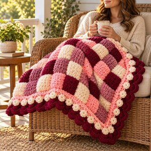 May include: A chunky crochet blanket featuring a checkered pattern in shades of cream, light pink, mauve, and deep burgundy. The blanket is finished with a decorative scalloped border in burgundy and small cream-colored floral accents.