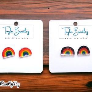 May include: Two pairs of rainbow earrings. One pair has a red outer edge and the other has a black outer edge. Both pairs are on white cards with the text "Taylor Brantley @BrilliantlyTay" printed on them.