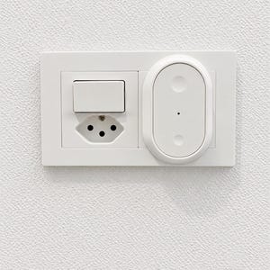 Peut inclure: Une plaque murale rectangulaire blanche comprenant un interrupteur, une prise électrique à trois broches et un dispositif de contrôle domotique ovale avec deux boutons circulaires et un petit voyant lumineux sur la droite.