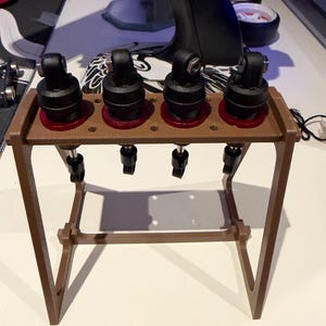 May include: A brown 3D-printed stand holding four black and red RC shock absorbers. The stand features a horizontal bar with four circular cutouts where the shocks are mounted, supported by two angled side legs and a base frame.