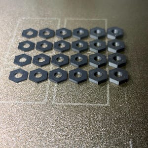 May include: A collection of twenty-four small, black, hexagonal-shaped plastic nuts arranged in a grid pattern. Each individual nut features a circular hole in the centre and a flat, matte surface texture, consistent with 3D printed components.