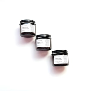 Op de afbeelding: Drie kleine, donkerbruine glazen potten met zwarte deksels zijn diagonaal op een witte achtergrond geplaatst. Elke pot heeft een wit etiket met zwarte tekst: "PINK BLOOM Organic Facial Mask". De potten zijn voor huidverzorging.