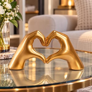 May include: A gold-colored sculpture featuring two hands positioned to form a heart shape. The sculpture has a smooth, reflective metallic finish and is designed to stand upright on a flat surface, showcasing a symmetrical hand gesture.