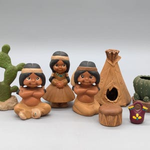 May include: A collection of small ceramic figurines including two seated figures, one standing figure, a teepee, a cactus, a potted plant, a drum, and a small purple vessel. The figures have dark hair, headbands, and tan skin tones.