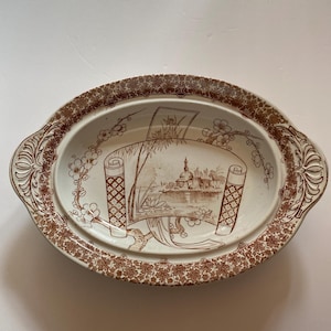 May include: An oval ceramic serving dish with a brown transferware pattern. The center features a landscape illustration framed by a scroll, surrounded by floral motifs. The rim has a dense floral border and two decorative handles.