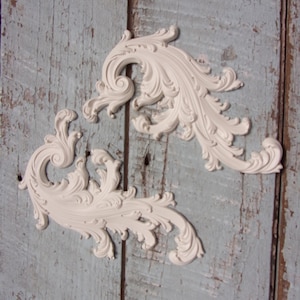 May include: Two off-white, ornate decorative scrollwork pieces. The scrollwork features a detailed, swirling leaf and vine design. The pieces are set against a weathered, light blue wooden background.