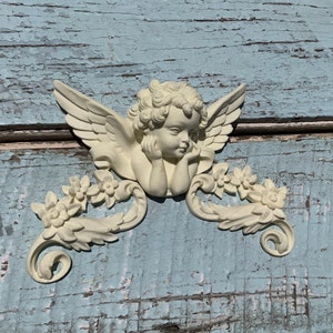 May include: A white ceramic cherub figurine with wings and floral accents. The cherub is holding its chin and has a sweet expression. The figurine is mounted on a blue wooden background.