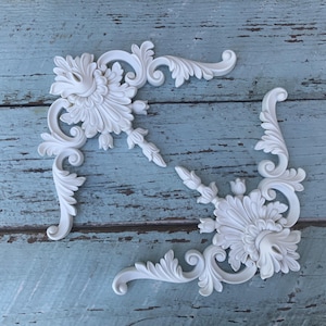 May include: Two white, ornate corner appliques featuring intricate acanthus leaf carvings and floral motifs. These decorative pieces have a symmetrical, scrolling design with detailed relief work, showcasing a classic, elaborate pattern in a matte finish.