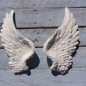 Puede incluir: Un par de alas de &aacute;ngel blancas decorativas con un dise&ntilde;o de plumas. Las alas est&aacute;n montadas en una superficie de madera azul claro.