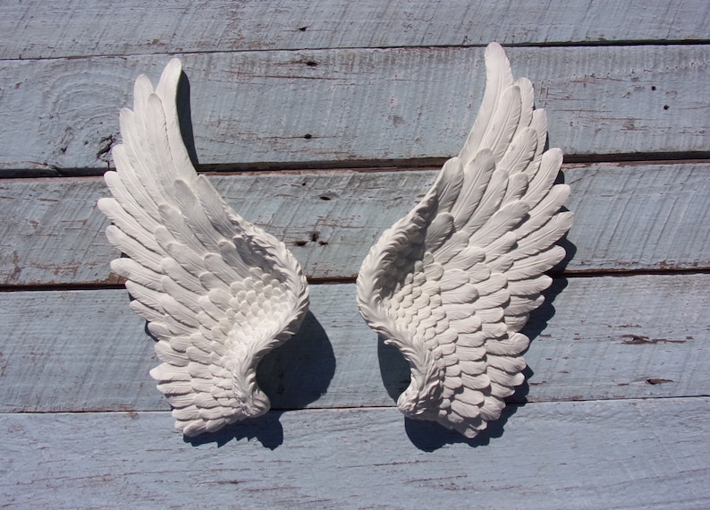 Puede incluir: Un par de alas de &aacute;ngel blancas decorativas con un dise&ntilde;o de plumas. Las alas est&aacute;n montadas en una superficie de madera azul claro.
