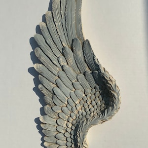 Puede incluir: Un ala de &aacute;ngel decorativa, grande, gris y con un acabado desgastado. El ala est&aacute; hecha de un material texturizado y tiene un dise&ntilde;o de plumas realista.