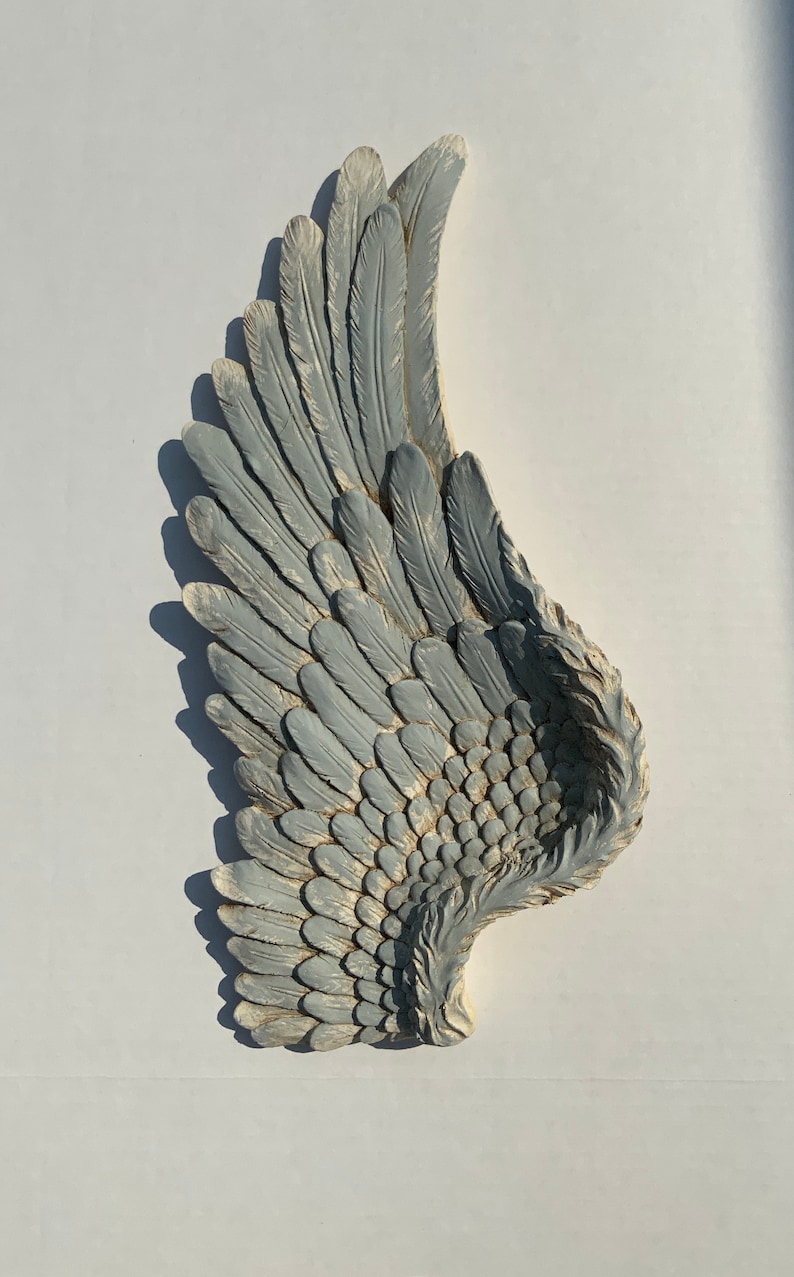 Puede incluir: Un ala de &aacute;ngel decorativa, grande, gris y con un acabado desgastado. El ala est&aacute; hecha de un material texturizado y tiene un dise&ntilde;o de plumas realista.