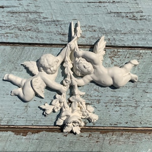 May include: Two white cherub figurines holding onto a floral design with a white background. The cherubs are holding onto a branch of flowers with their hands.