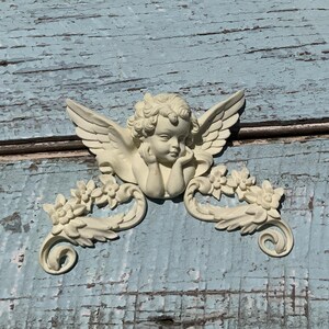 May include: A white cherub figurine with wings and floral accents. The cherub is holding its chin and looking down. The figurine is mounted on a weathered blue wooden surface.