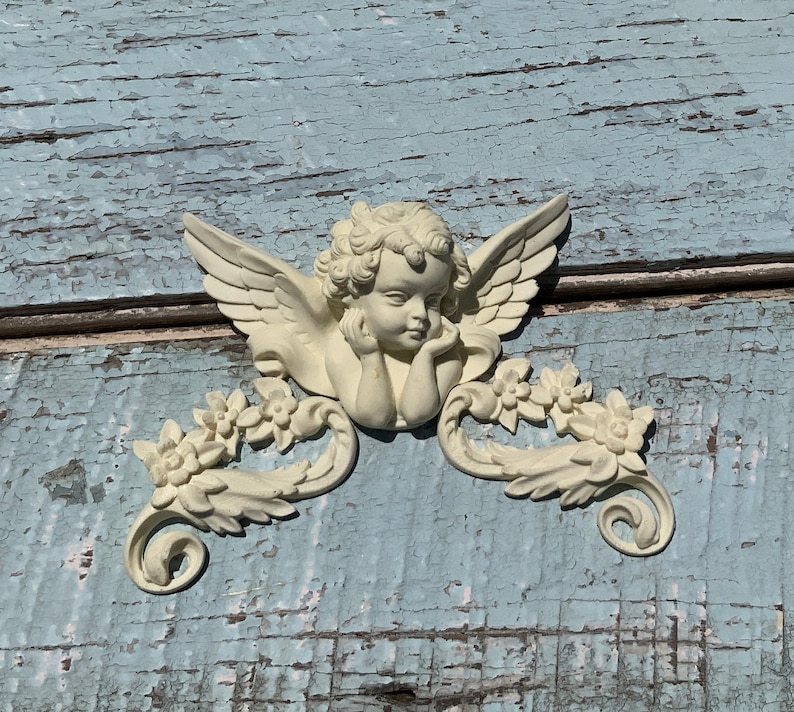 May include: A white cherub figurine with wings and floral accents. The cherub is holding its chin and looking down. The figurine is mounted on a weathered blue wooden surface.