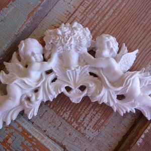 May include: White ceramic ornament featuring two cherubs holding a basket of flowers. The cherubs are sculpted in a delicate, whimsical style and are surrounded by ornate floral details.