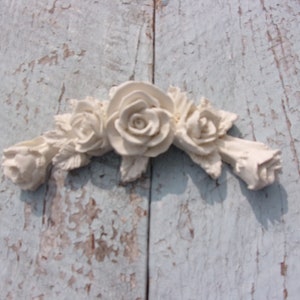 May include: White ceramic rose garland with five roses and leaves. The garland is attached to a light blue wooden surface.