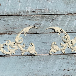 May include: Two white ornate decorative wall accents with a scroll design. The accents are made of a lightweight material and have a distressed finish.