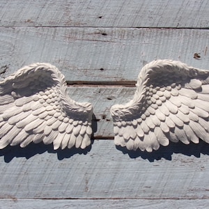 Puede incluir: Un par de alas de &aacute;ngel blancas decorativas con textura de plumas. Las alas est&aacute;n montadas en una superficie de madera azul claro.