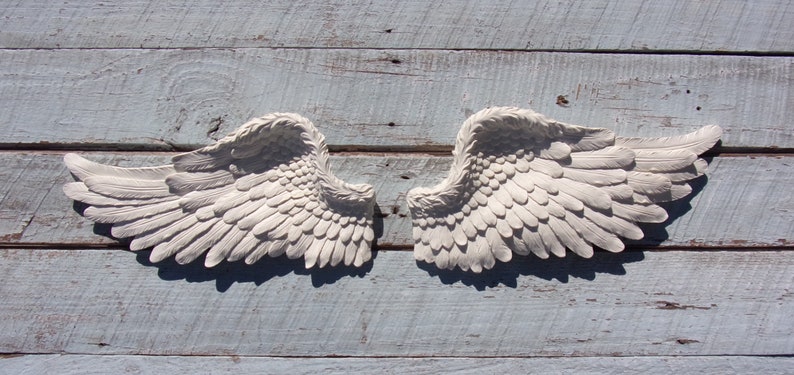 Puede incluir: Un par de alas de &aacute;ngel blancas decorativas con textura de plumas. Las alas est&aacute;n montadas en una superficie de madera azul claro.