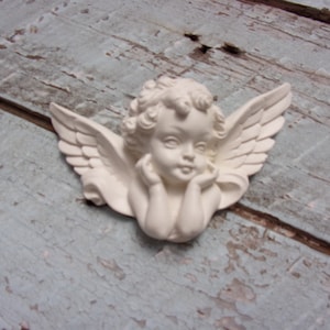 May include: White ceramic cherub figurine with wings and a sweet expression, holding its hands together under its chin. The figurine is attached to a light blue wooden surface.