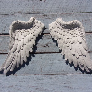 Puede incluir: Un par de alas de &aacute;ngel blancas decorativas con textura de plumas. Las alas est&aacute;n montadas en una superficie de madera azul claro.