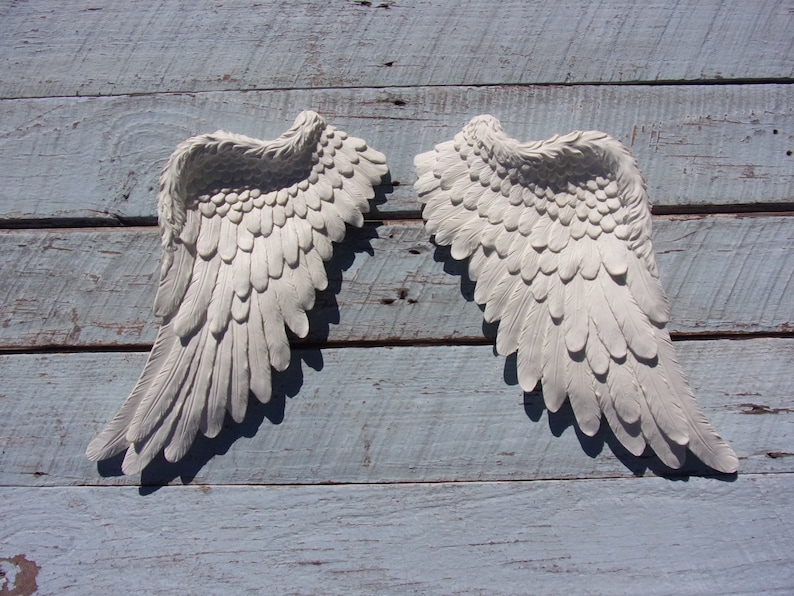 Puede incluir: Un par de alas de &aacute;ngel blancas decorativas con textura de plumas. Las alas est&aacute;n montadas en una superficie de madera azul claro.