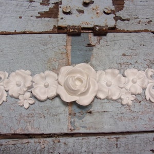 May include: White decorative floral trim with a large rose in the center and smaller flowers on either side. The trim is made of a lightweight material and has a delicate, ornate design.