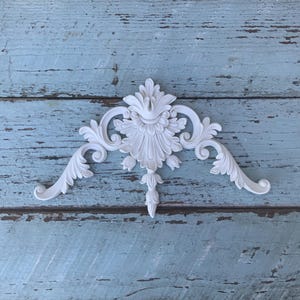 May include: A white decorative architectural applique featuring a symmetrical floral and acanthus leaf scrollwork design. The piece has a central pointed element with radiating petals and curved side extensions that taper to rounded ends.
