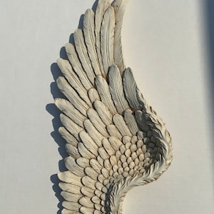 Puede incluir: Un ala de &aacute;ngel de yeso blanco con un acabado desgastado. El ala est&aacute; esculpida con plumas detalladas.