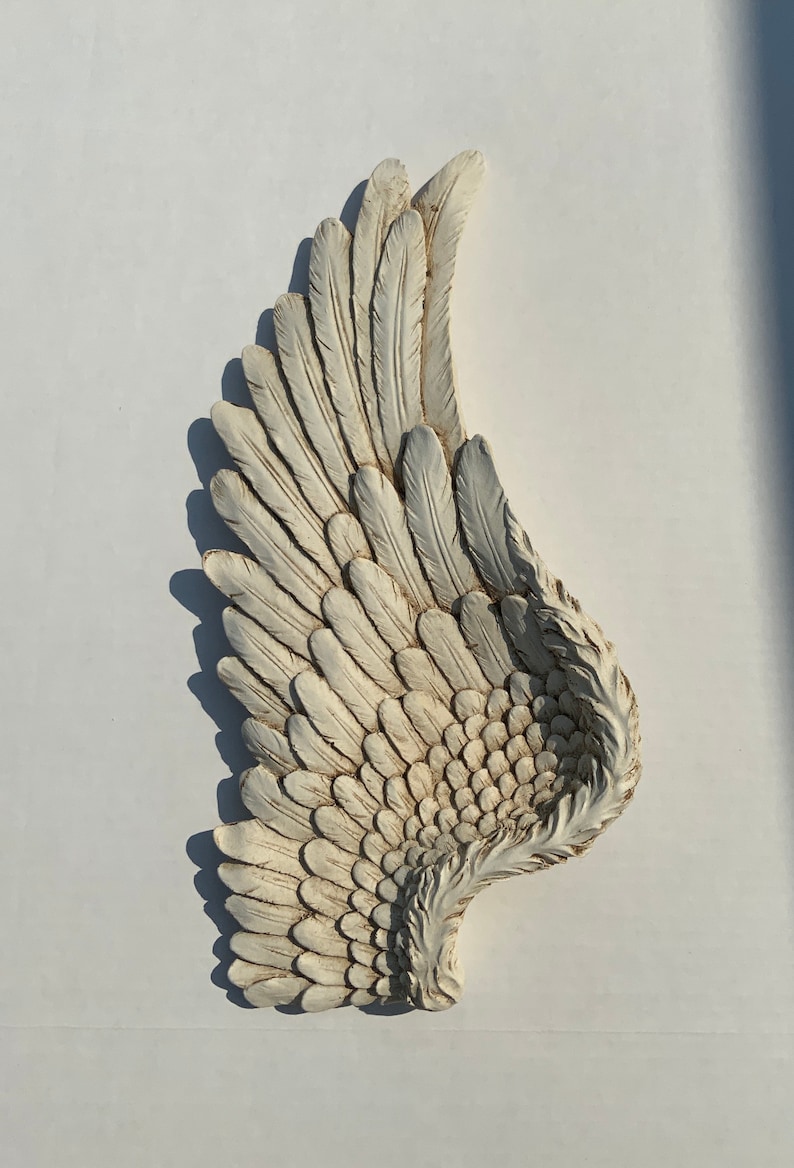 Puede incluir: Un ala de &aacute;ngel de yeso blanco con un acabado desgastado. El ala est&aacute; esculpida con plumas detalladas.