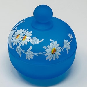 May include: A round, blue trinket box with a matching lid and a small knob. The box is decorated with white daisies with yellow centers and green leaves. The box is made of a matte material.