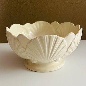 May include: A cream-colored ceramic bowl featuring a scalloped rim and a molded exterior design that mimics the appearance of overlapping seashells. The bowl is supported by a circular pedestal base and has a smooth, glossy finish throughout.