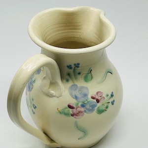 May include: Cream-colored ceramic pitcher with a rounded body and a curved handle. The pitcher is decorated with hand-painted blue and pink floral designs and green leaves. The pitcher has a spout for easy pouring.
