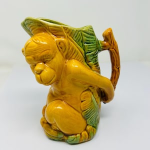 May include: A ceramic pitcher shaped like a monkey sitting on a green leaf. The pitcher has a brown handle and is glazed in yellow and green.