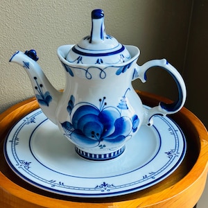 May include: White ceramic teapot with blue floral designs and a matching saucer. The teapot features a curved handle, a spout with a decorative element, and a lid with a finial. The blue and white color scheme gives it a classic look.