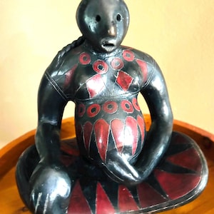 May include: A black ceramic figurine of a seated person with red and black painted details. The figure has a braided hairstyle and is seated with legs crossed. The figurine is on a wooden surface.