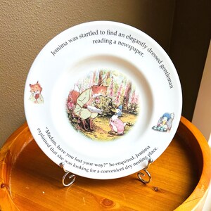 May include: White ceramic plate featuring a colorful illustration of a fox and a duck in a forest scene. The plate has text around the rim, including the phrase "Jemima was startled to find an elegantly dressed gentleman reading a newspaper."