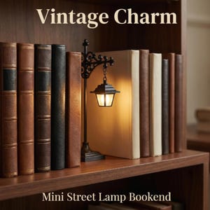 Op de afbeelding: Een zwarte metalen mini straatlantaarn boekensteun met een gloeiend licht, geplaatst tussen verschillende in leer gebonden boeken op een houten plank. De tekst 'Vintage Charm' en 'Mini Street Lamp Bookend' is zichtbaar.