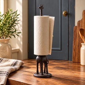 May include: A dark brown cast iron paper towel holder featuring a giraffe figure. The giraffe's long neck serves as the central spindle for a roll of white paper towels, while its four legs provide a stable base on a circular platform.