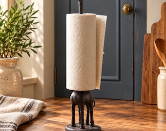 May include: A dark brown cast iron paper towel holder featuring a giraffe figure. The giraffe's long neck serves as the central spindle for a roll of white paper towels, while its four legs provide a stable base on a circular platform.