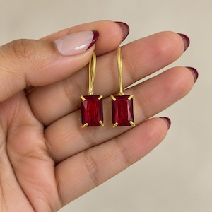 Puede incluir: Un par de pendientes colgantes con piedras rectangulares de color rojo intenso facetadas, engastadas en garras de metal dorado. Cada pendiente cuelga de un sencillo gancho dorado, mostrando una piedra preciosa roja clara y vibrante.