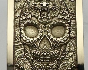 Puede incluir: Una barra de metal rectangular de tono plateado con un diseño en relieve de un cráneo humano coronado por una cabeza de tigre gruñendo. La superficie está decorada con intrincados patrones florales y motivos de rosas que rodean la figura central.