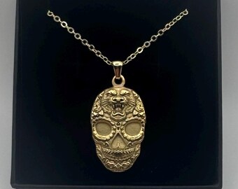 Puede incluir: Un collar con colgante en tono dorado que presenta un diseño de calavera con una cabeza de tigre rugiente integrada en la parte superior. La calavera está decorada con intrincados grabados florales y cuelga de una fina cadena.