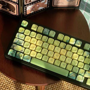 May include: A black mechanical keyboard featuring custom keycaps with a gradient of yellow and green hues. Several keys display intricate landscape illustrations, while the standard QWERTY layout is printed in a dark font on the keycaps.